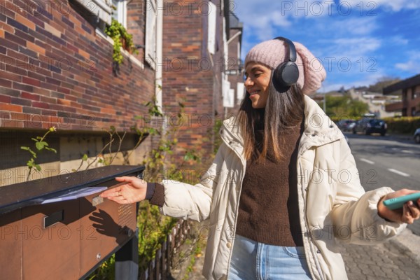 Young woman smiling, wearing a beanie and headphones, mailing a letter into an outdoor mailbox on an urban street, holding a smartphone in her hand during a sunny day