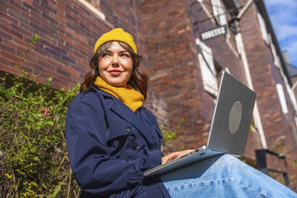 Woman wearing a warm yellow beanie and trench coat, smiling while sitting outdoors with a laptop on her lap, enjoying remote work in an urban setting on a sunny day