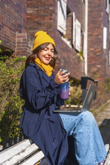 Young woman sitting on a park bench in an urban environment, using her laptop for digital nomad remote work and staying hydrated with a sports water bottle