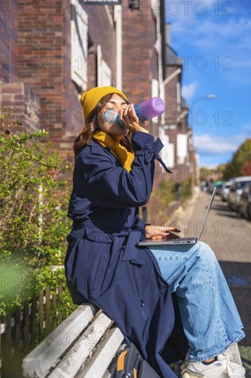 Woman hydrating with a water bottle while working remotely on her laptop, sitting on a bench in an urban setting during an autumn day, symbolizing flexible work and connection