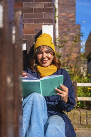 Young woman in a yellow beanie sits on a bench against a brick wall, smiling and reading a book while enjoying warm autumn sunlight in a relaxed urban setting