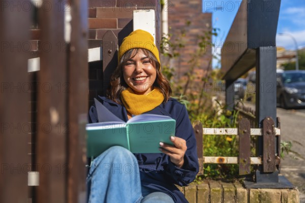 Young woman smiling in a cozy beanie and scarf, sitting on a city street enjoying a sunny autumn day while relaxing and reading a book, calm and content