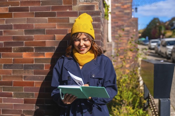 Young woman dressed in a trench coat and yellow beanie enjoying journaling and creative writing in her notebook during a sunny autumn day on an urban street
