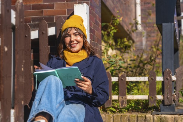 Young woman wearing a yellow beanie and scarf. Sitting outdoors in autumn sunlight. Smiling while reading a journal and looking at the camera. Capturing a moment of personal reflection and relaxation