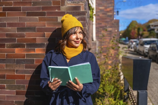 Young woman wearing a yellow beanie and scarf, holding an open journal while standing against a brick wall on an autumn day, looking away thoughtfully