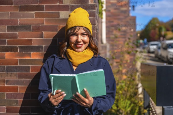 Young woman wearing a yellow beanie and scarf, dressed in a trench coat, standing outdoors in warm sunny autumn light, reading an open book against a brick wall