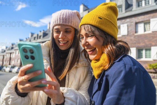 Two young women, dressed in winter hats and warm clothing, are laughing while looking at a mobile phone together on a bright day in an urban setting, sharing a cheerful moment