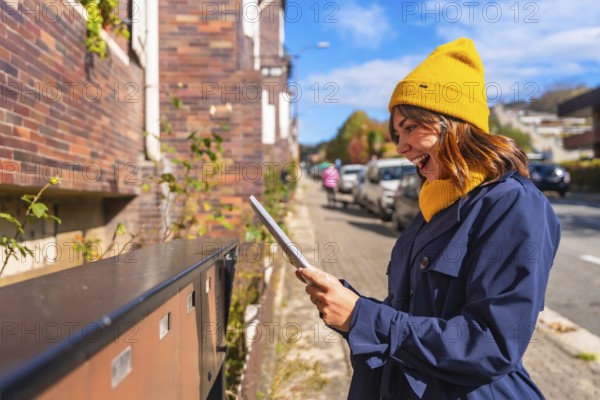Young smiling woman wearing a yellow beanie and blue trench coat showing excitement finding a letter in an outdoor street mailbox, illustrating communication, surprise, and positive emotion