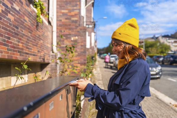 Woman wearing a yellow beanie and blue trench coat, smiling while sending a letter through a street mailbox, connecting with others through traditional mail in an urban setting
