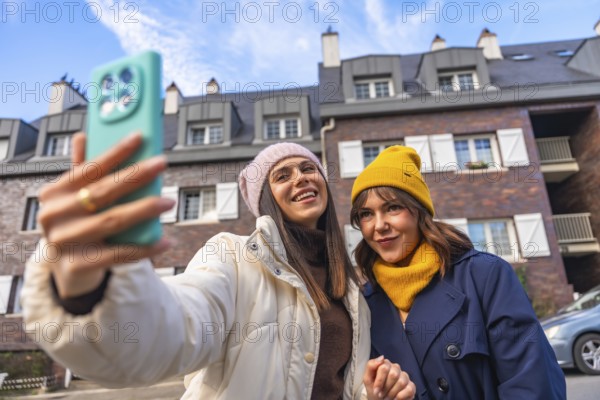 Two young women friends enjoying a fun moment together, smiling and taking a selfie outdoors in a city street during winter, wearing warm beanies and coats