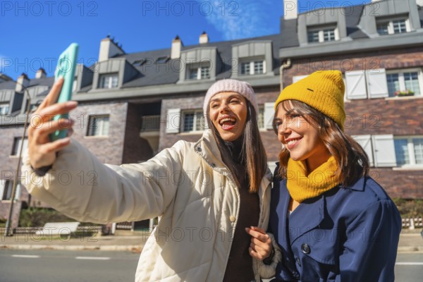 Two cheerful young women friends are enjoying a sunny winter day in the city, smiling and taking a fun selfie together with a smartphone, sharing a moment of joyful friendship
