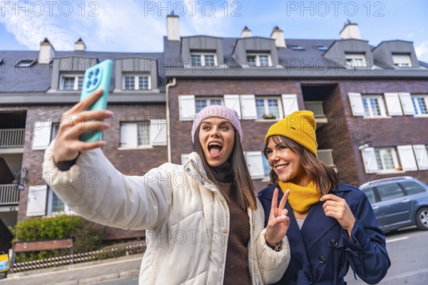 Two cheerful young women take a sunny winter selfie on a city street, bundled in beanies and jackets, smiling and capturing a fun travel moment together