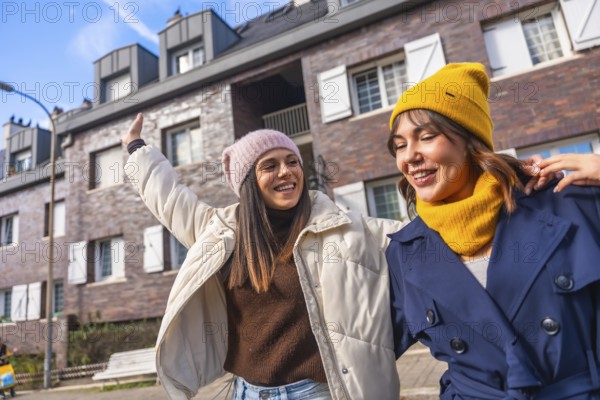 Two young women friends walking a city street in winter coats and beanies, laughing and chatting together, enjoying sunny urban weekend freedom and warm friendship vibes