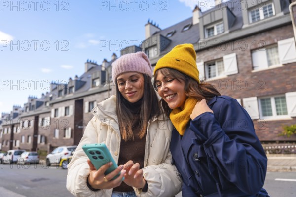 Two young women in winter coats and beanies smile and share a phone while walking a city street, enjoying a candid moment of friendship, tech and urban exploration