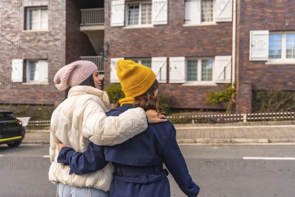Two young women in warm winter clothes walk arm in arm down a city street, smiling and enjoying a cheerful, cozy moment of friendship and togetherness outdoors