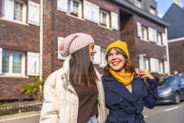 Young women friends walking down a sunny city street in warm winter fashion, smiling and chatting as they enjoy a cheerful, casual urban outing together