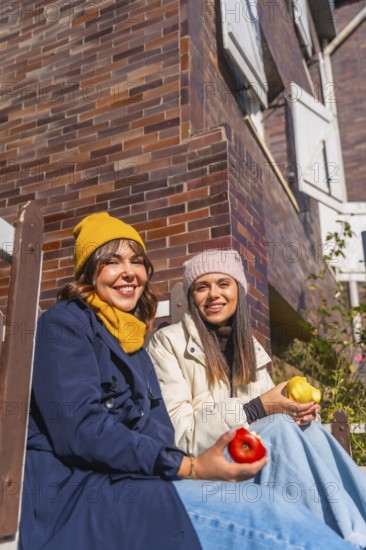 Two friends in warm winter clothes sit against a brick wall, smiling and sharing red and yellow apples on a sunny day, enjoying a casual, healthy moment together
