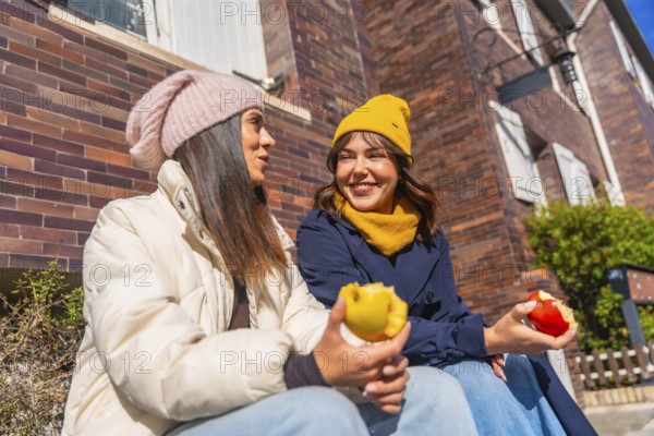 Two young women friends sitting together on a sunny autumn day, enjoying fresh apples while having a friendly conversation in an urban setting, representing friendship and healthy living