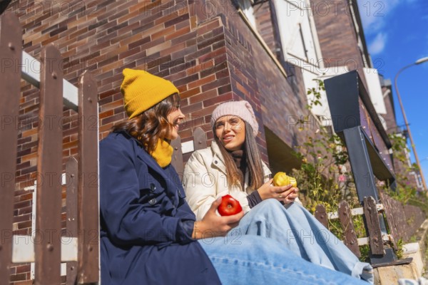 Two young women sit on outdoor steps by a brick wall under blue sky, smiling and chatting while enjoying apples, cozy in beanies and coats on a sunny autumn day