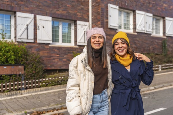 Two smiling young women friends are standing together on an urban street in front of a brick building, dressed in warm stylish clothing, enjoying a sunny autumn day