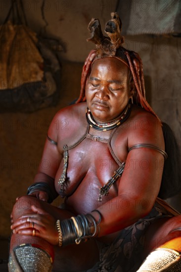 Elderly Himba woman rubbing herself with red paste for skin care, in a hut, traditional Himba village, Kaokoveld, Kunene, Namibia