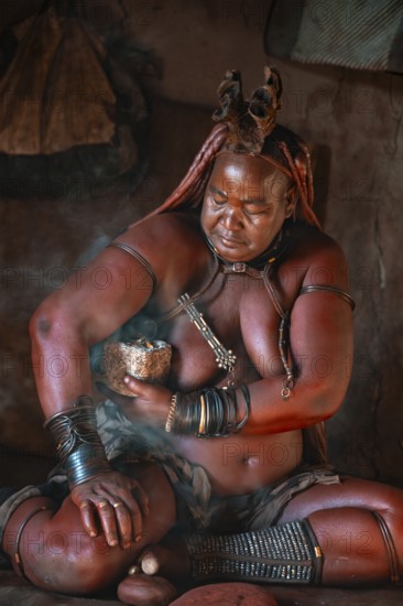 Natural perfume, elderly Himba woman smokes her hair for good smell, in a hut, traditional Himba village, Kaokoveld, Kunene, Namibia