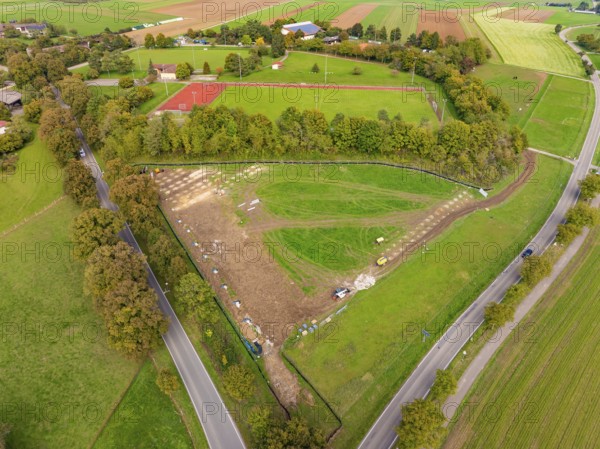 Green landscape with tennis court and roads flanked by trees under cloudy sky, PV Freifaechenanlage construction, Weil der Stadt, Germany