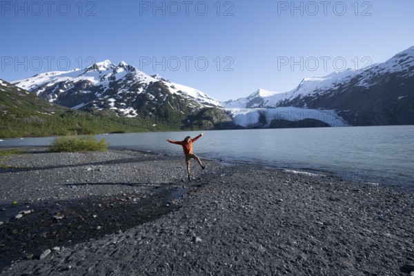 Young man jumping on a pebble beach on a glacial lake, Snowy Mountains and Portage Glacier on Portage Lake glacial lake, Chugach National Forest, Alaska, USA