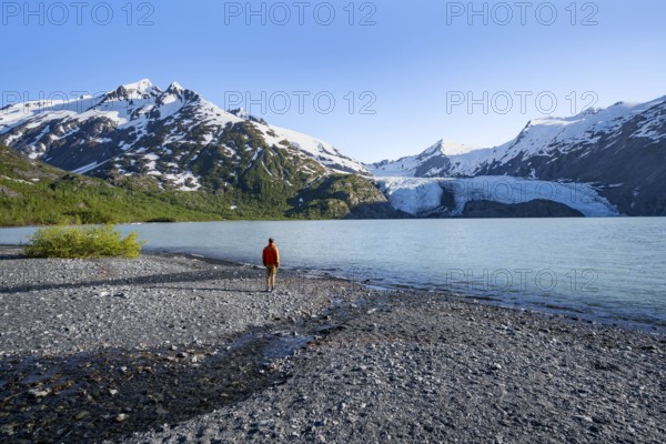 Young man on a pebble beach on a glacial lake, snowy mountains and Portage Glacier glacier on Portage Lake glacial lake, Chugach National Forest, Alaska, USA