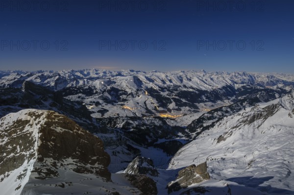 Night shot, long exposure from 2506 meter high SÃ¤ntis to the wintery, snow-covered Swiss Alps with the illuminated village of Wildhaus, during a clear full moon night, Canton of Appenzell, Switzerland