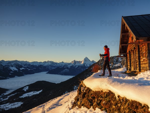 Mountaineers enjoy the sunrise in front of an alpine hut in winter, the Watzmann behind, fog in the valley, Berchtesgaden Alps, Scheibenkaser, Marktschellenberg, Berchtesgadener Land, Upper Bavaria, Bavaria, Germany