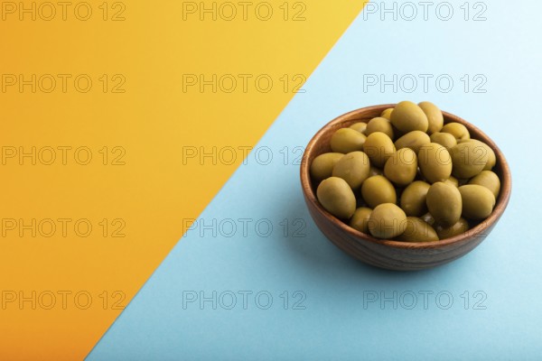 Fresh green olives in wooden bowl on blue and orange pastel paper background. side view, copy space