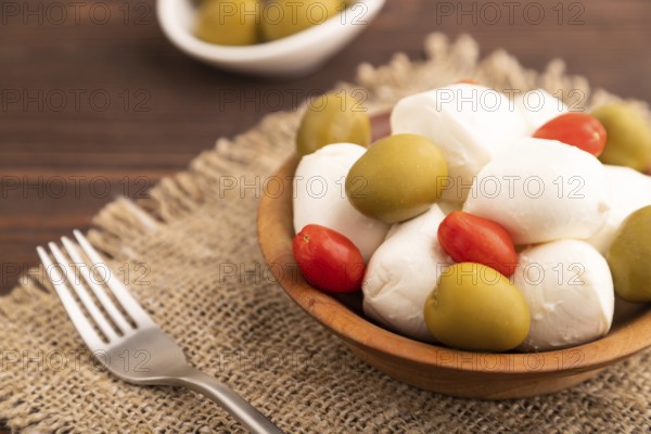 White Mozzarella cheese, with tomatoes and olives in wooden bowl on brown wooden background and linen textile, side view, close up, selective focus