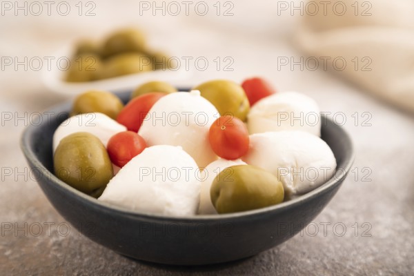 White Mozzarella cheese, with tomatoes and olives in blue bowl on brown concrete background and linen textile, side view, close up, selective focus