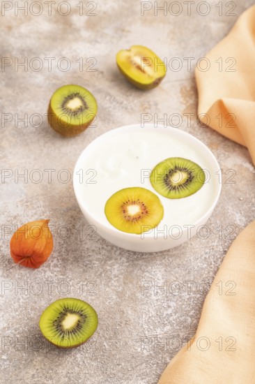 Yogurt with kiwi in white bowl on brown concrete background and orange linen textile, side view, close up, minimalism