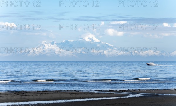 View over Cook Inlet to white mountain peaks of Mount Iliamna, motor boat rides on the ocean, mountains of the Aleutian Range, Anchor Point, Anchor River State Recreation Area, Alaska, USA