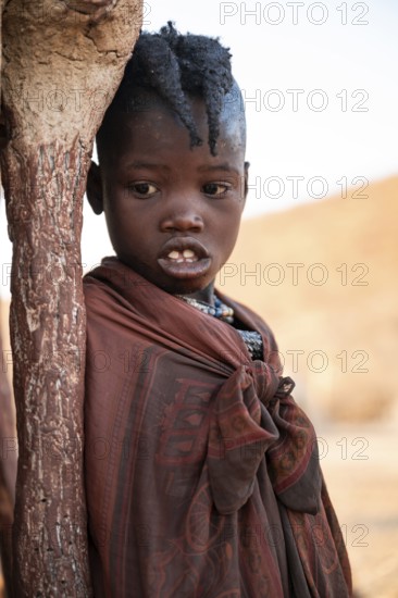 Himba girl leaning at a traditional clay hut, in the morning, traditional Himba village, Kaokoveld, Kunene, Namibia