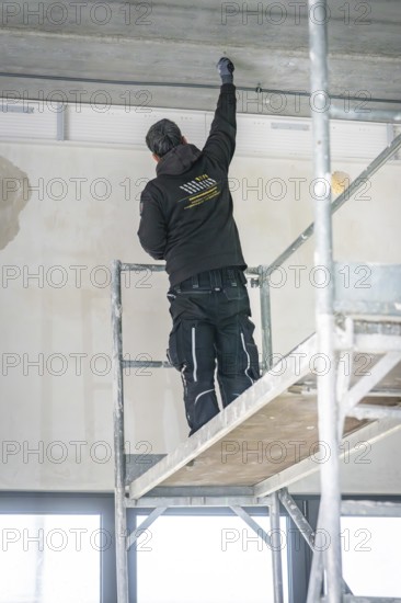 A worker stands on scaffolding and works on the ceiling in a renovated room, electrician, new building, Germany