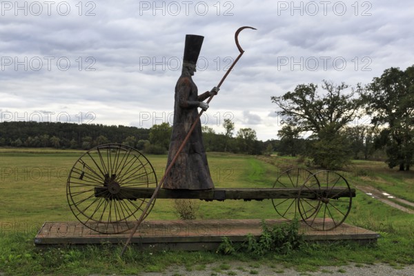 Sculpture in a meadow, traditional grim reaper, figure carrying a scythe, standing on cart with large wheels, symbol of death, sculpture of ferryman Charon by Bernd Streiter on the Elbe cycle path, twilight, Mödlich, Prignitz, Brandenburg, Germany