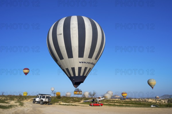 Large striped hot air balloon lands next to jeep and red car in clear landscape, Göreme Tarihî Millî Park, Nevsehir Province, Nevsehir, Cappadocia, Capadocia, Cappadocia, Central Anatolia, Turkey