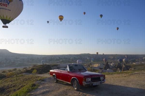 A red convertible stands in front of a vast landscape with hot air balloons in the morning sky, Ford, Göreme Tarihî Millî ParkÄ±, Nevsehir Province, Nevsehir, Cappadocia, Capadocia, Cappadocia, Central Anatolia, Turkey