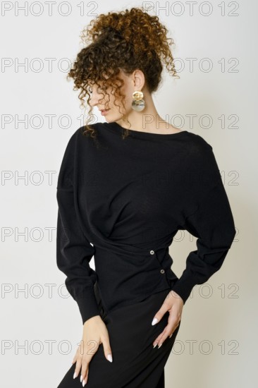 A stylish model with curly hair poses against a neutral backdrop. She wears black blouse with twist and accentuates her look with bold accessories. The overall vibe is chic and modern