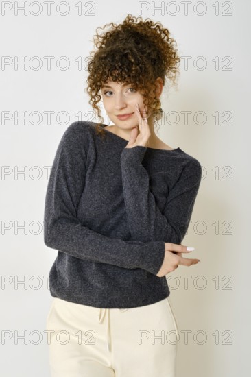 Cheerful woman in dark sweater and light pants standing against white wall with a playful expression