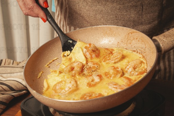 A person stirs shrimp in a creamy sauce in a frying pan on the stove, showcasing the cooking process in a warm kitchen filled with natural light