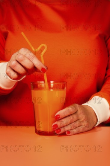 A woman in an orange sweater, stirring with a straw, orange juice in a glass, orange background
