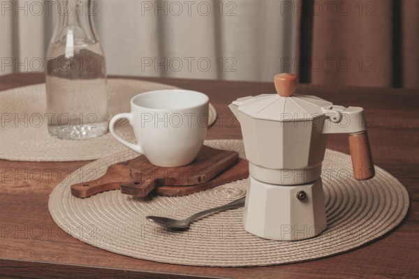 A moka pot sits on a wooden cutting board beside a white cup on a round placemat. A glass bottle of water is in the background, capturing a cozy morning scene