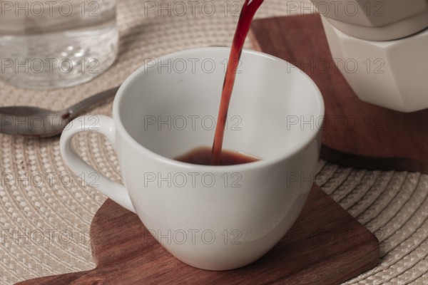Hot coffee is poured into a white cup resting on a wooden coaster. A serene breakfast setting is visible, inviting warmth and comfort in the morning light