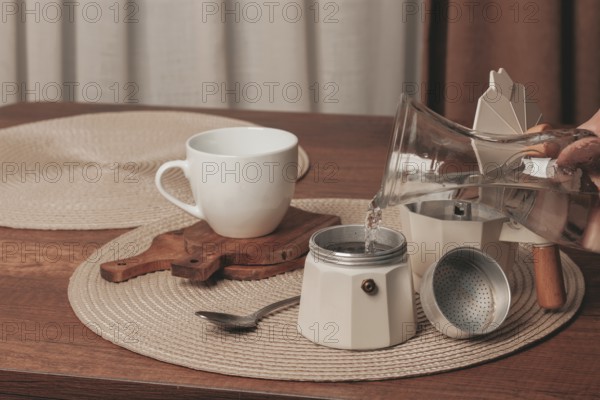 Hot water is being poured from a glass kettle into a coffee maker on a wooden table. A white cup awaits the fresh brew in a simple kitchen environment