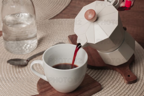 Freshly brewed coffee is being poured from a stovetop espresso maker into a white cup on a wooden table. A clear glass water bottle sits nearby, adding to the warm, inviting setting