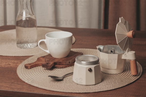 A cozy scene features a white cup resting on a wooden coaster next to a moka pot and a glass water bottle. Soft morning light fills the room, creating a warm atmosphere